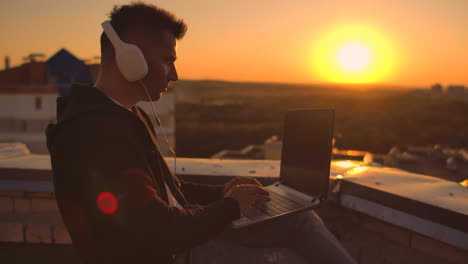 Ein-Mann-Mit-Kapuzenpullover-Sitzt-Auf-Dem-Dach-Und-Hört-Musik-Mit-Kopfhörern-Und-Tippt-Auf-Der-Tastatur-Eines-Laptops.-Laufen-Bei-Sonnenuntergang.-Freiberufler-Arbeitet-Bei-Sonnenuntergang-Mit-Einem-Laptop